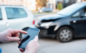 Eine Person wählt auf dem Smartphone, im Hintergrund ist ein Auffahrunfall zu sehen.